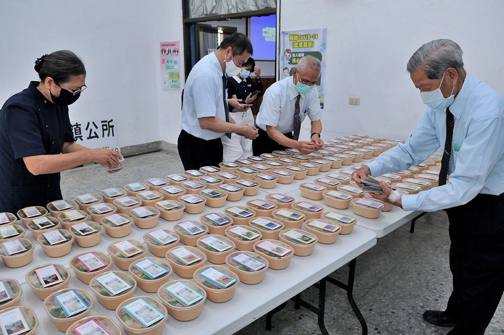 85歲的慈濟志工賴秋約(右)幫忙將兩張小卡片放在素食便當上面,讓公所同仁能夠QR CODE,上網點閱素食食譜。