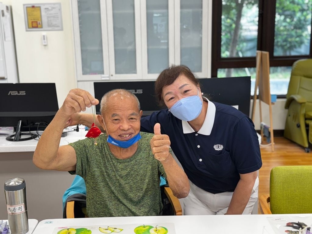陪伴長者的志工沈碧鳳(右)有感而發地說:「阿公阿嬤來到日照中心,不僅能和大家聊天互動,還能動手學東西、做健康操,生活多了很多樂趣。」